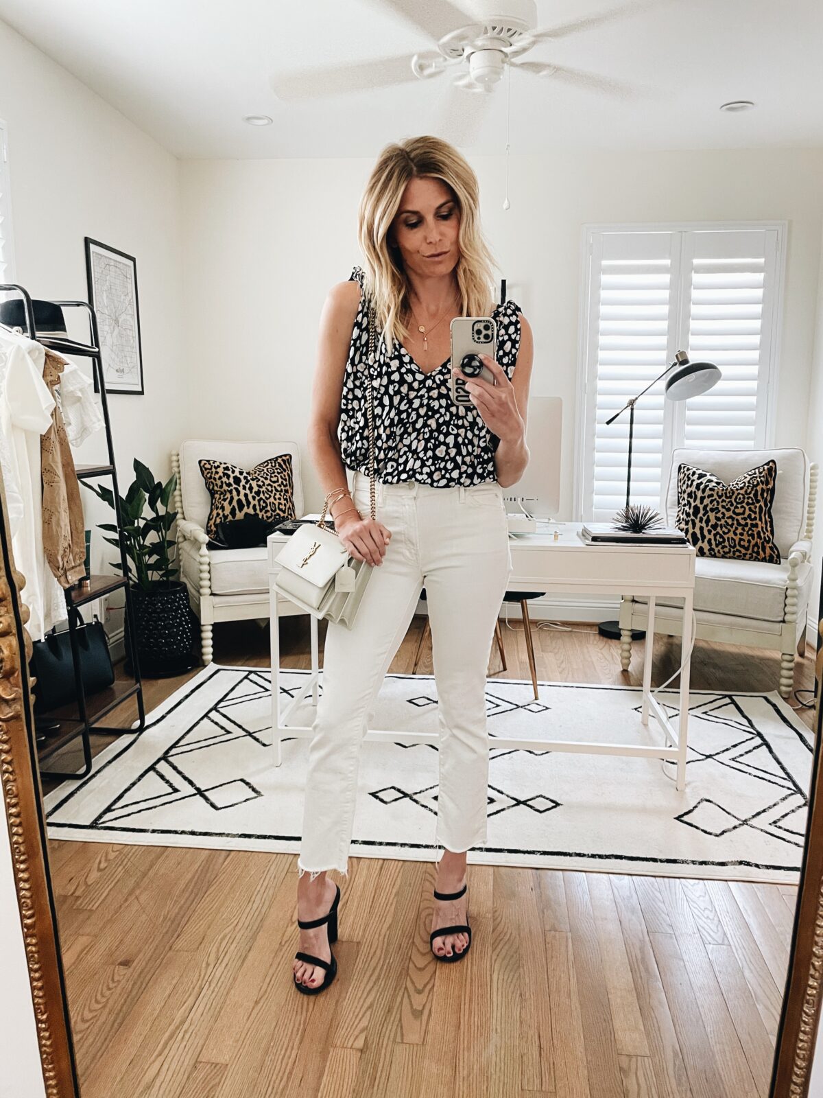 woman taking a selfie at home and wearing a printed top, white pants, and black high heels